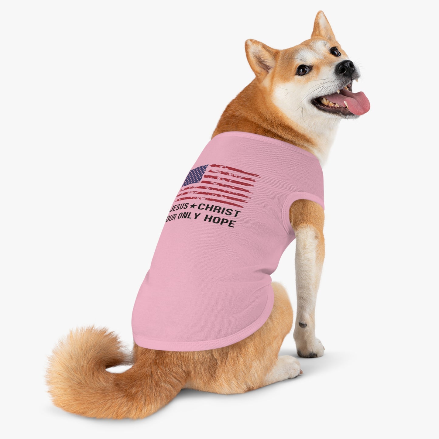 A Shiba Inu dog sitting on a white background wearing a pink sleeveless t-shirt. The shirt features a distressed American flag graphic and the text "JESUS * CHRIST OUR ONLY HOPE" in black.