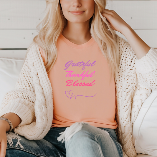 A photo of a woman sitting on a bed, wearing a peach-colored t-shirt with the words "Grateful," "Thankful," and "Blessed" printed in a gradient of pink and purple. She is looking at the camera and smiling.