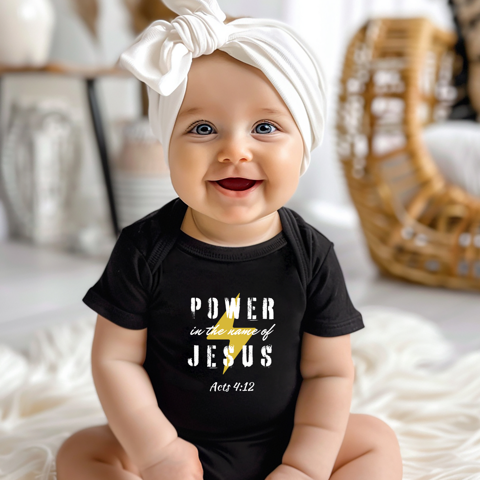A smiling baby with a white scarf tied around their head is wearing a black t-shirt with the words "POWER in the name of JESUS" and the Bible verse reference "Acts 4:12