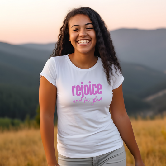 A photo of a young woman smiling and wearing a white t-shirt with the words "Rejoice and be glad" printed in pink. She is standing outdoors in a field with mountains in the background.